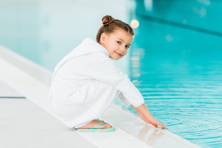 Cheerful Kid In Bathrobe Touching Water In Swimming Pool