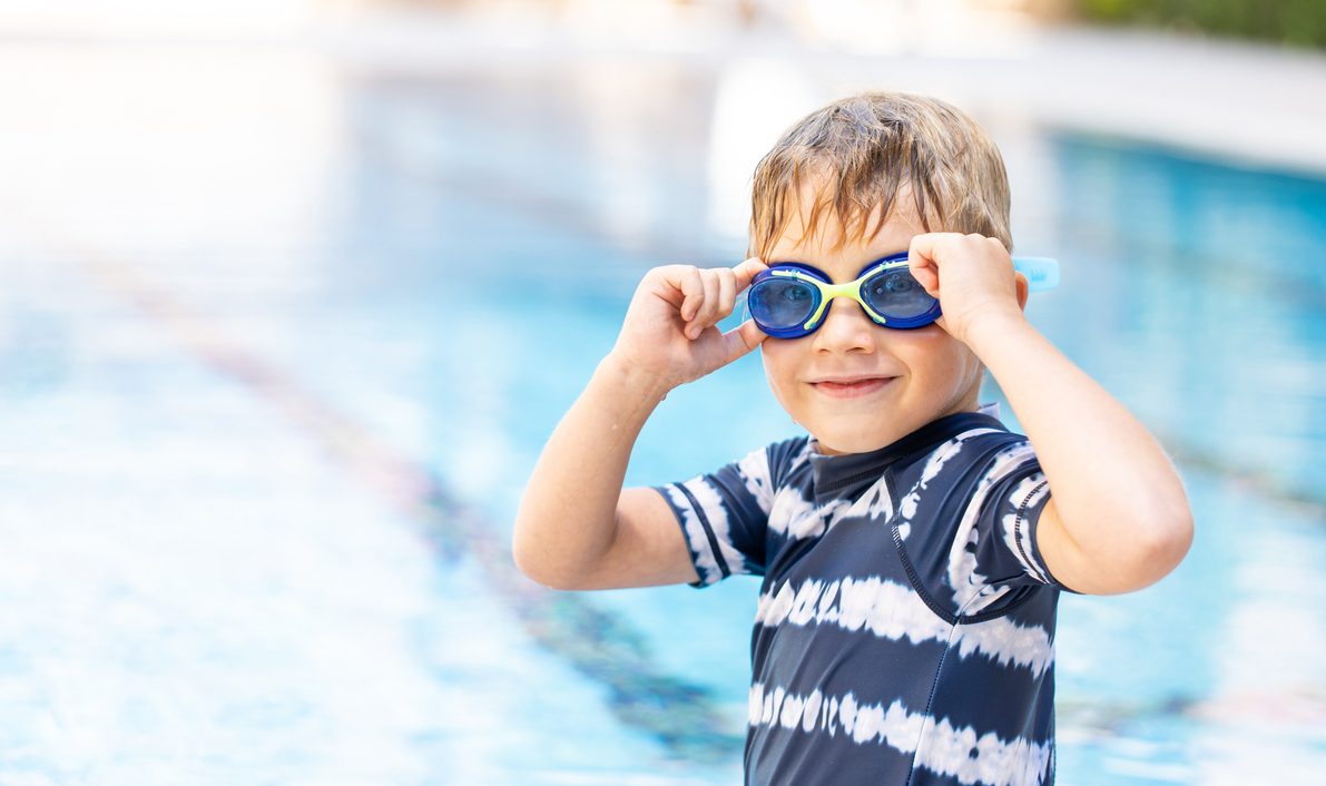 Portrait Of Active Kid In Swimming Goggles Sitting Near The Swimming Pool, Looking At Camera And Smiling